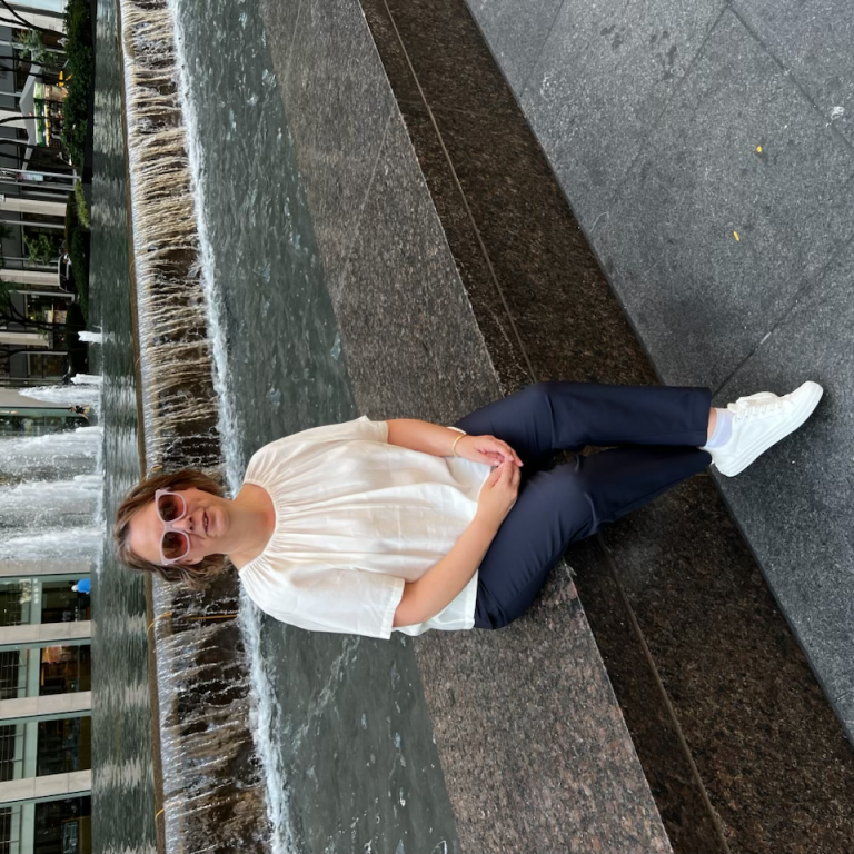Claire Banwell ER Co-Founder A woman sits by a fountain in an urban setting, wearing sunglasses and a light blouse.